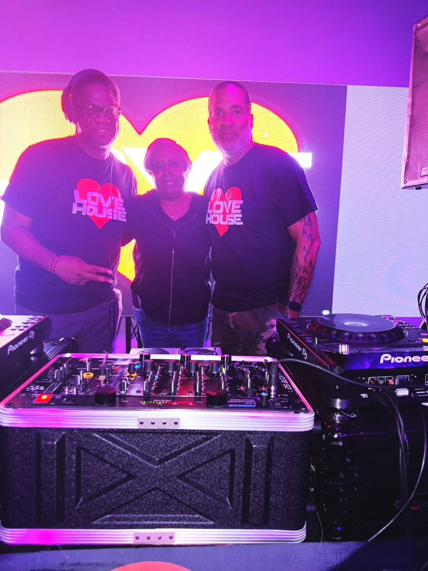 Three Love House crew members in branded tees pose behind Pioneer DJ gear and a flight case in front of a heart-lit screen.