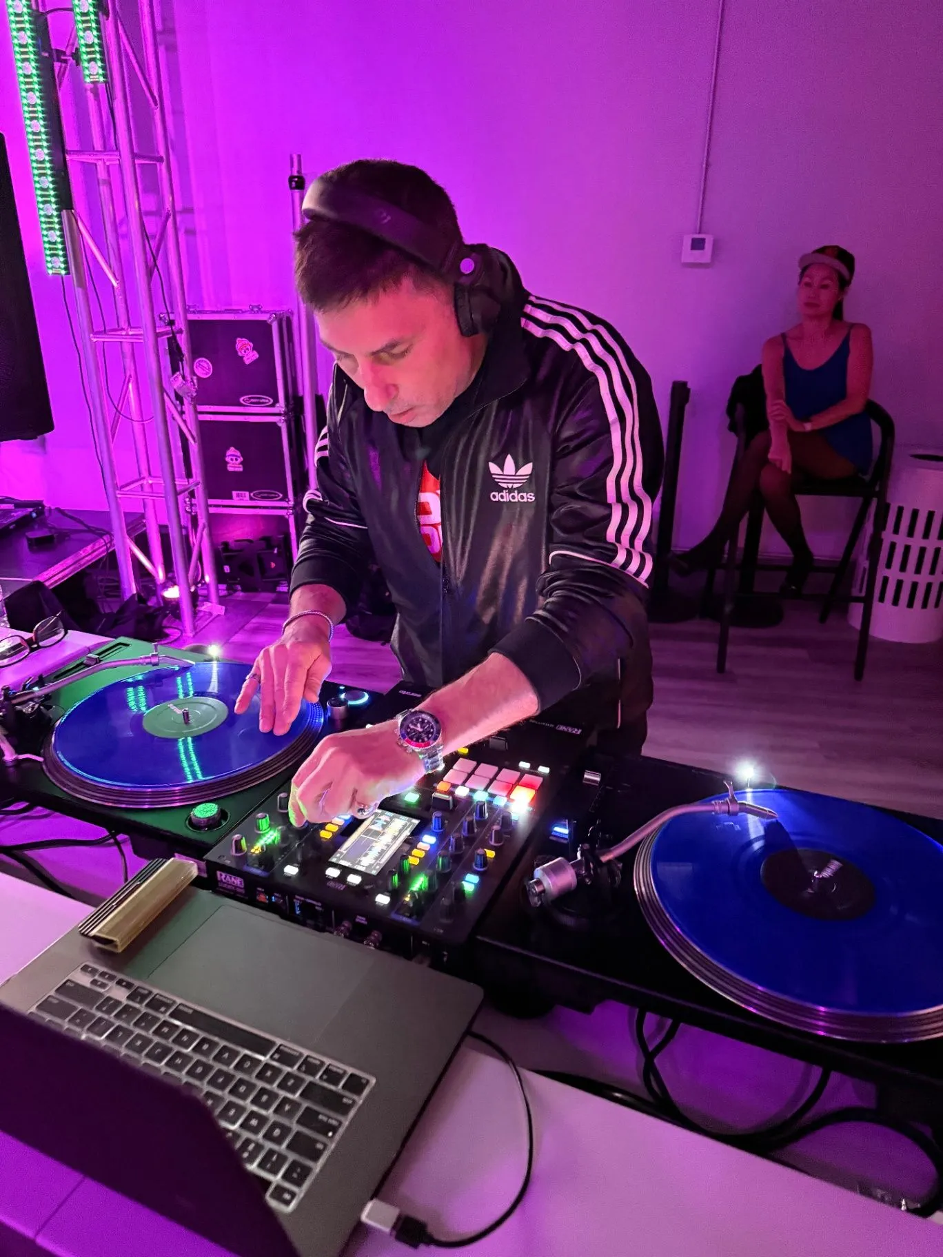 A DJ in an adidas track jacket and headphones mixes on two turntables with blue vinyl under purple and pink lighting.