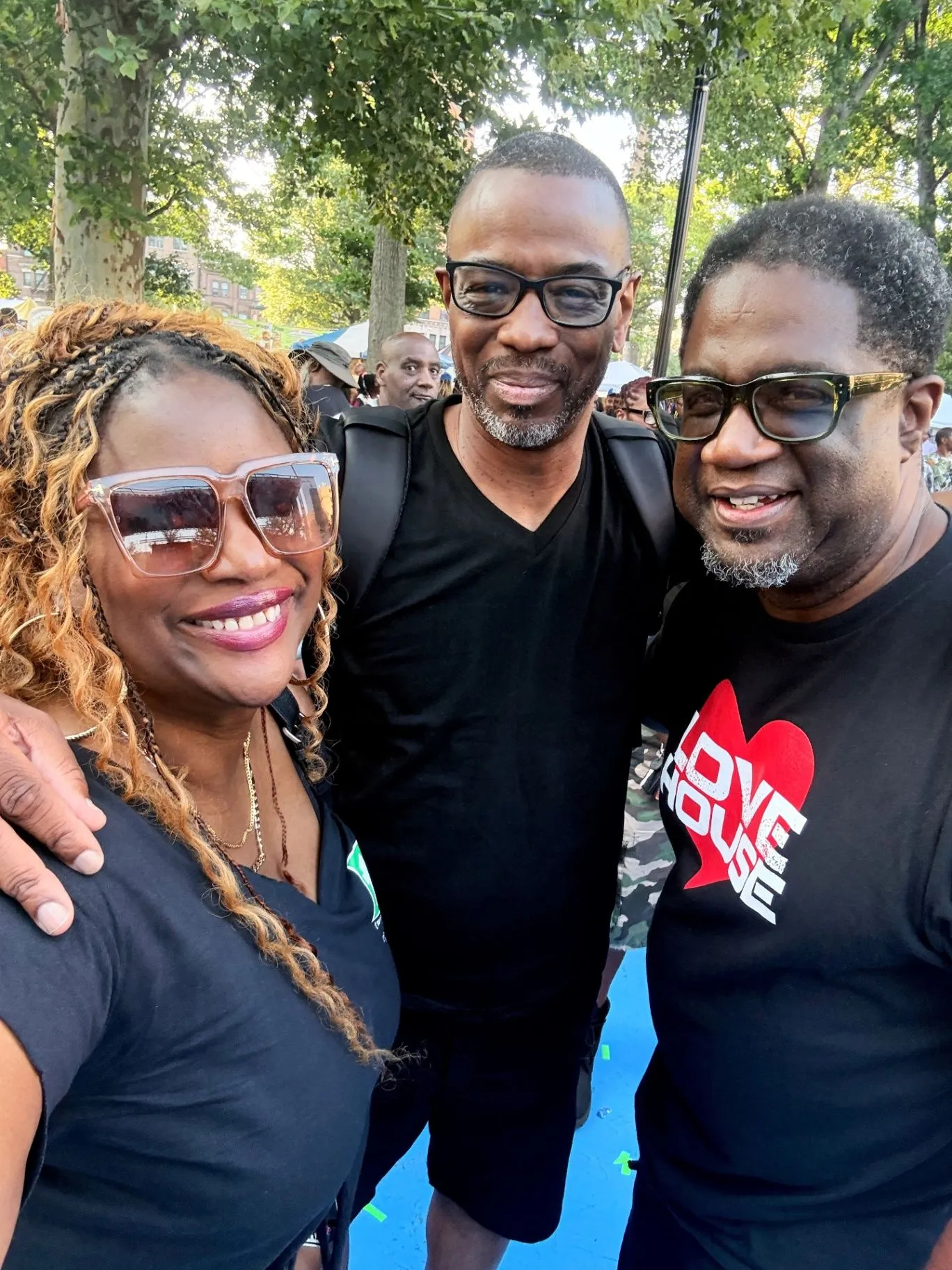 Three LoveHouse friends stand together outdoors in summer daylight at an event, with one wearing a Love House heart-logo tee.
