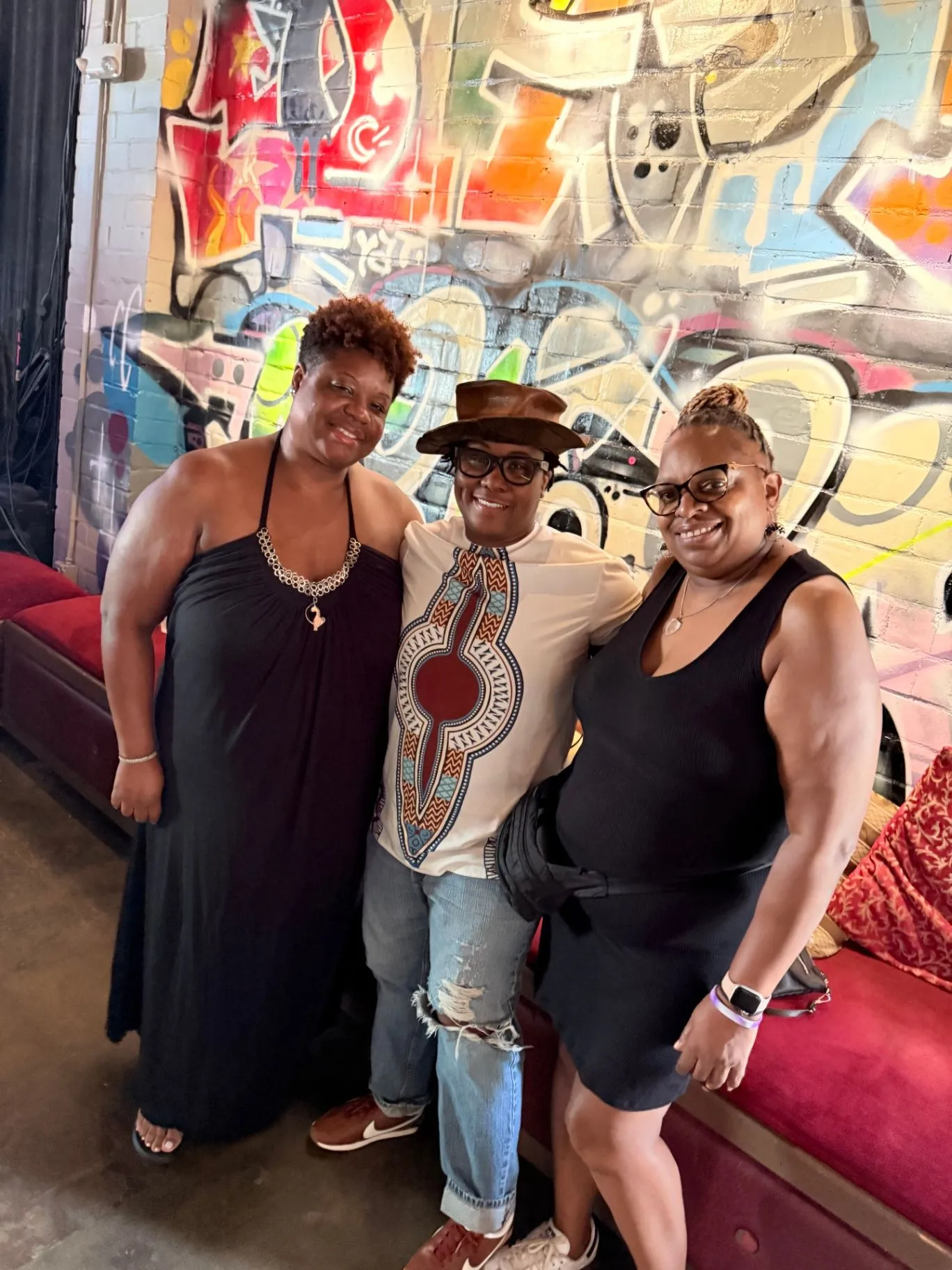 Three friends pose in front of a graffiti mural beside red lounge seating, with one wearing a patterned tunic and brimmed hat.