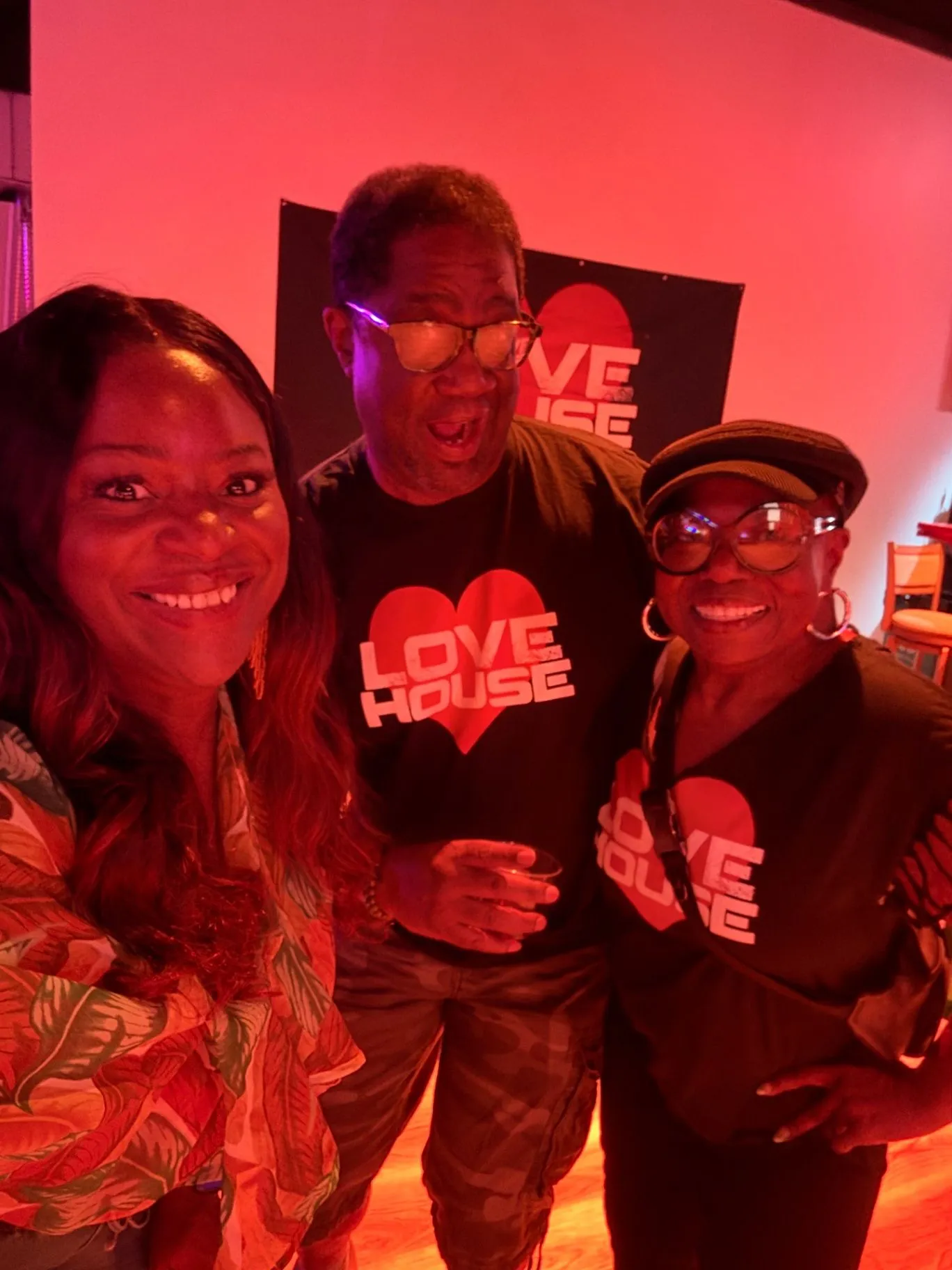 Three guests smile in warm red club lighting, with one wearing a Love House heart-logo tee and the Love House banner behind them.