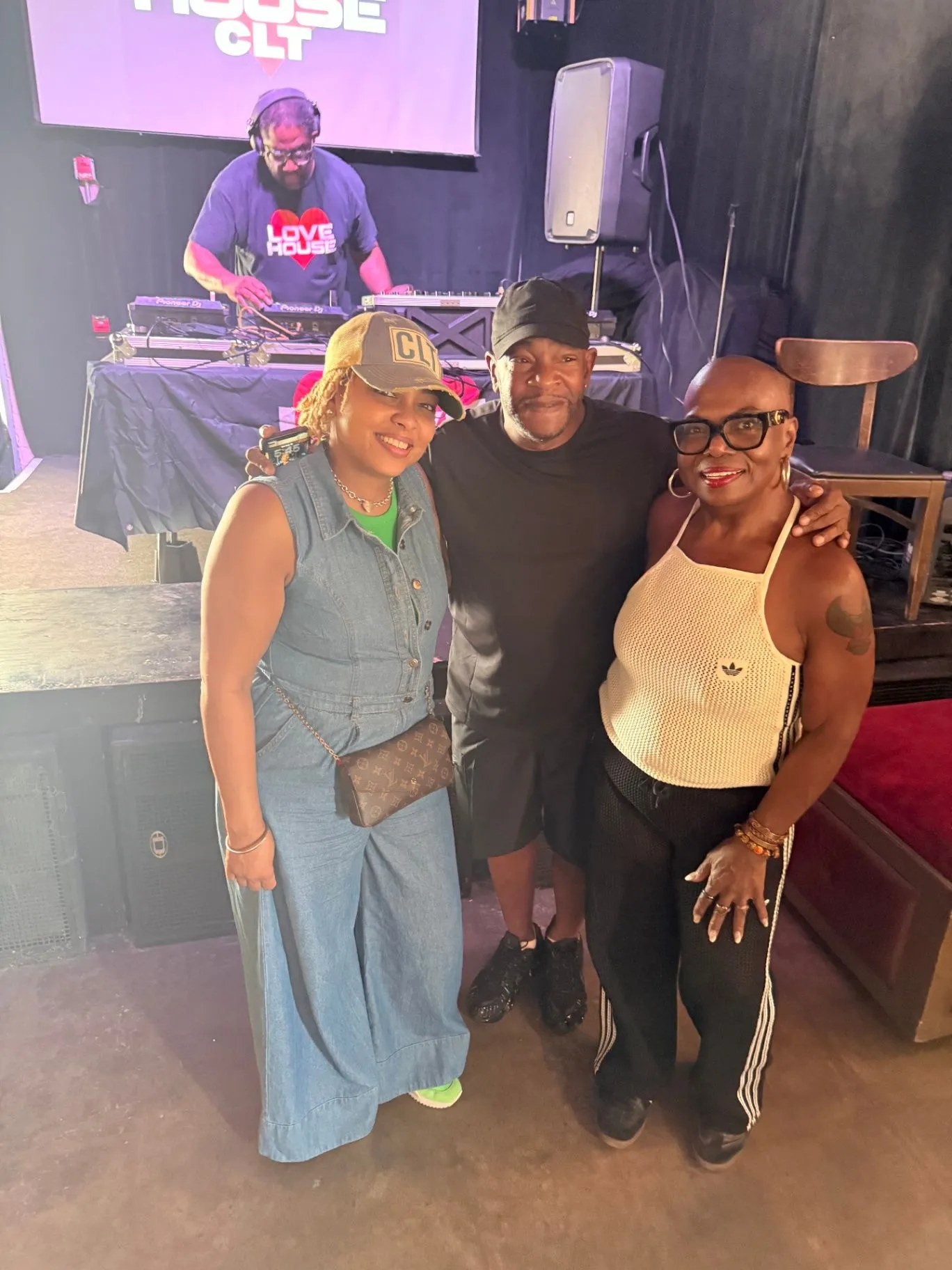 Three guests pose together in front of the stage at a LoveHouse CLT party, with a DJ in a Love House tee performing on Pioneer decks behind them and the LoveHouse CLT screen above.
