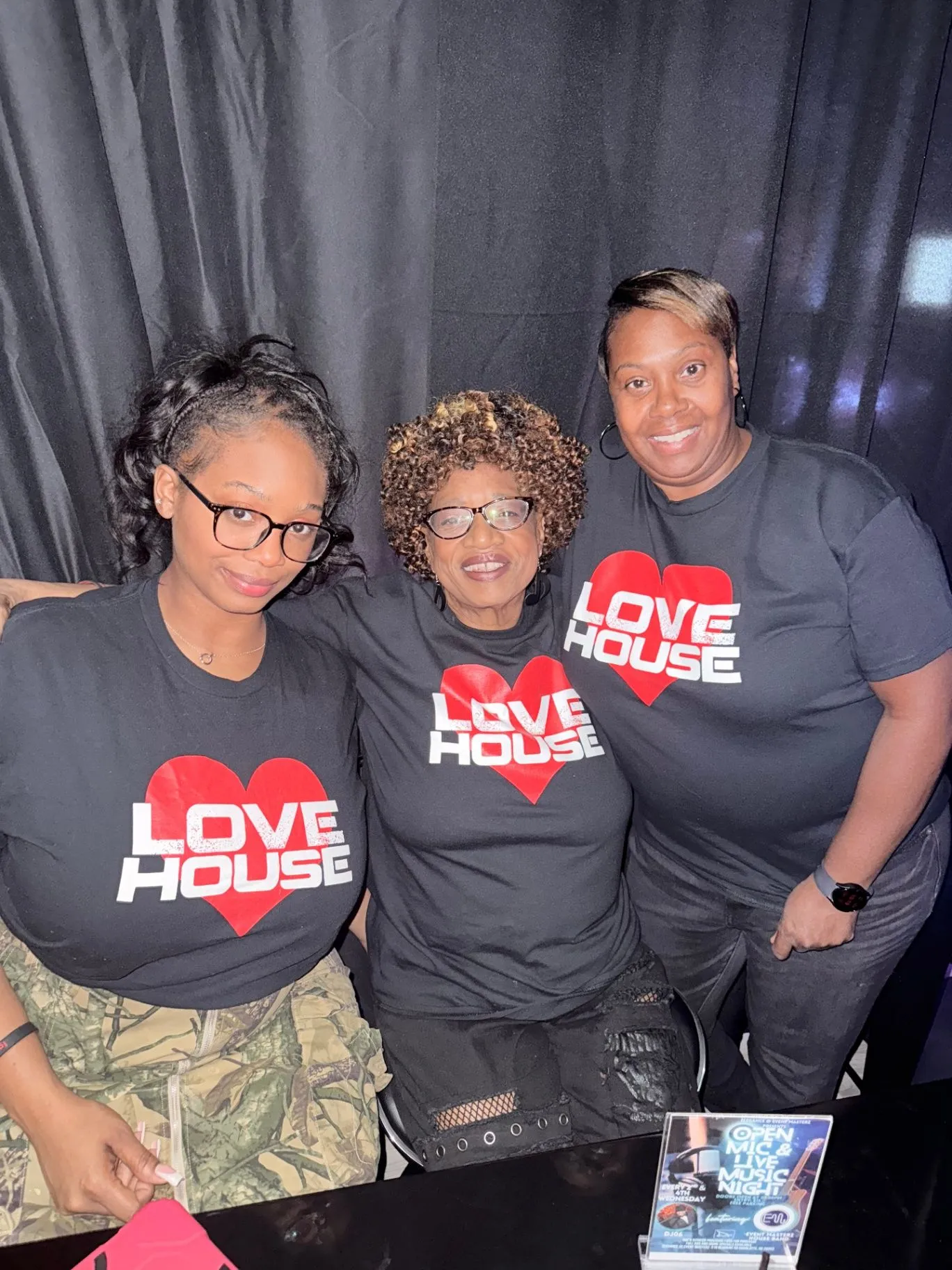 Three women in Love House heart-logo tees pose in front of a black curtain behind a table with an Open Mic & Live Music Night flyer.