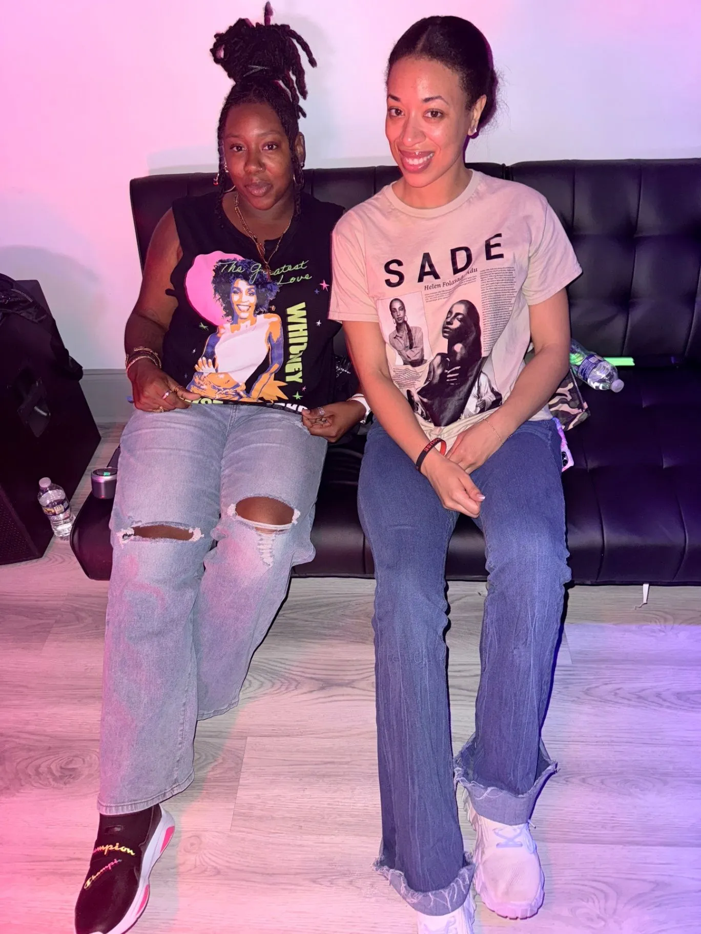 Two friends sit on a black leather sofa in pink lighting, wearing Sade and Whitney Houston graphic tees.